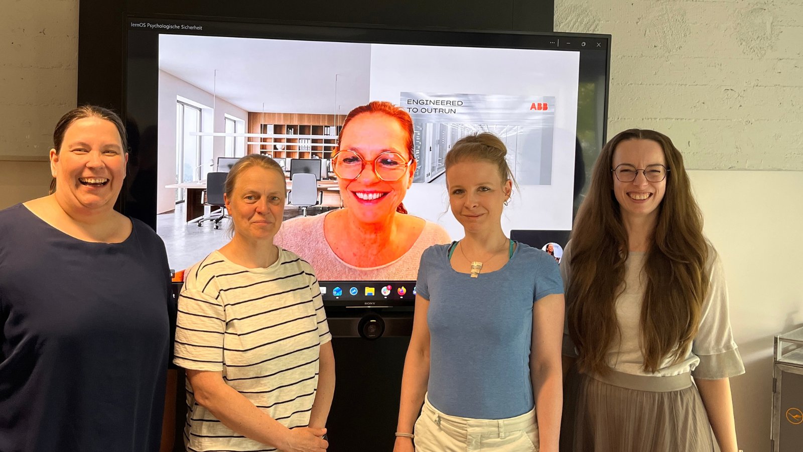 Vier Frauen stehen lächelnd vor einem großen Bildschirm, auf dem eine fünfte Frau per Videoschalte zu sehen ist