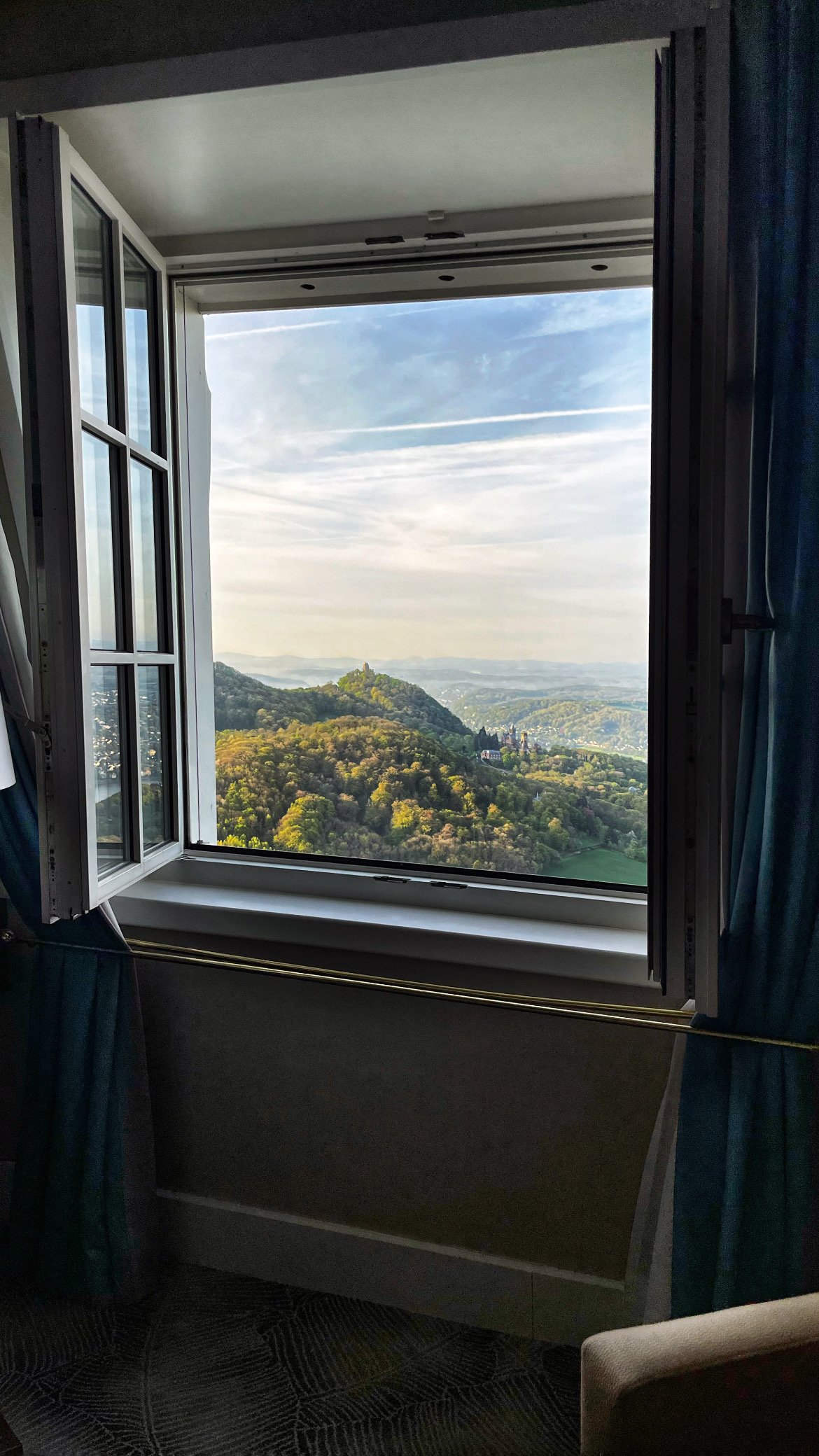 Blick aus einem geöffneten Hotelfenster auf bewaldete Hügel mit einer Burg im Morgenlicht, links und rechts hängen blaue Vorhänge