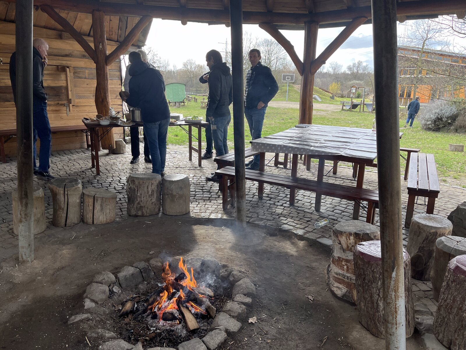 Covered fire pit with tree stumps as seats, men standing at a table with food in the background