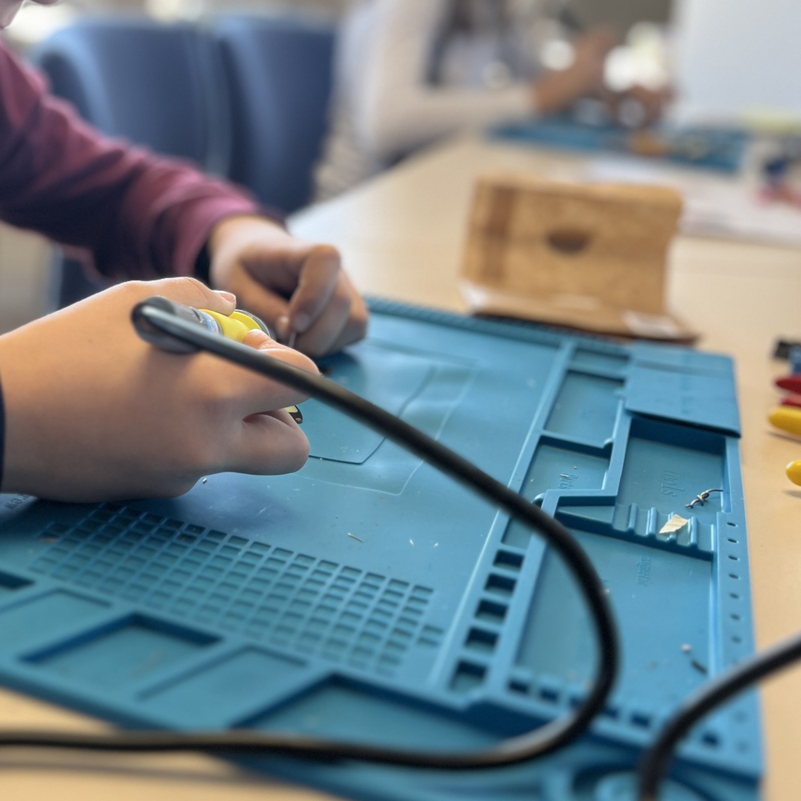 Soldering Hands hold a soldering iron over a blue silicone mat, other kids are working in the background