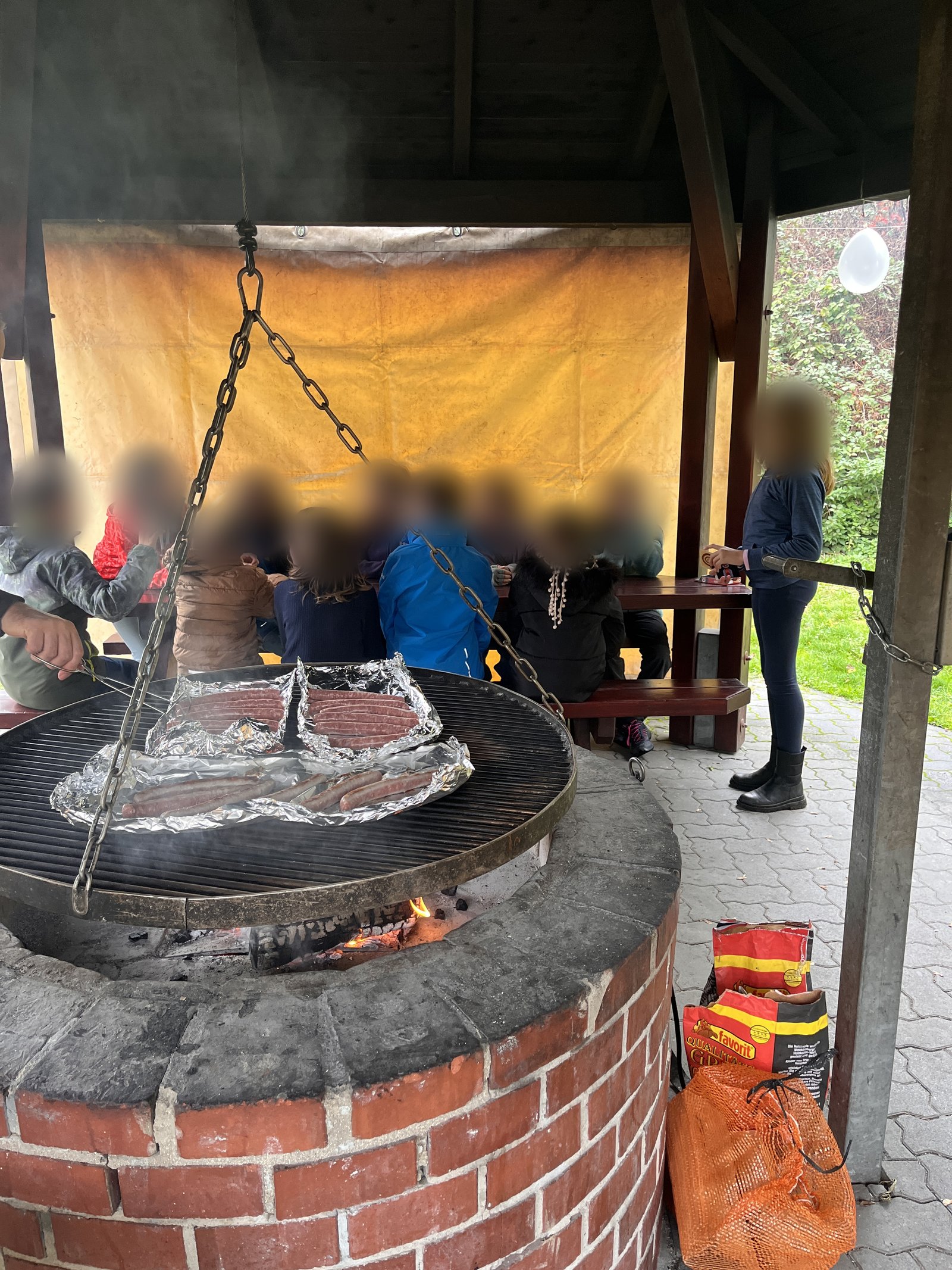 Children sit at a bench in a pavilion, in the foreground a brick grill with sausages on the swing grate. The faces are blurred.