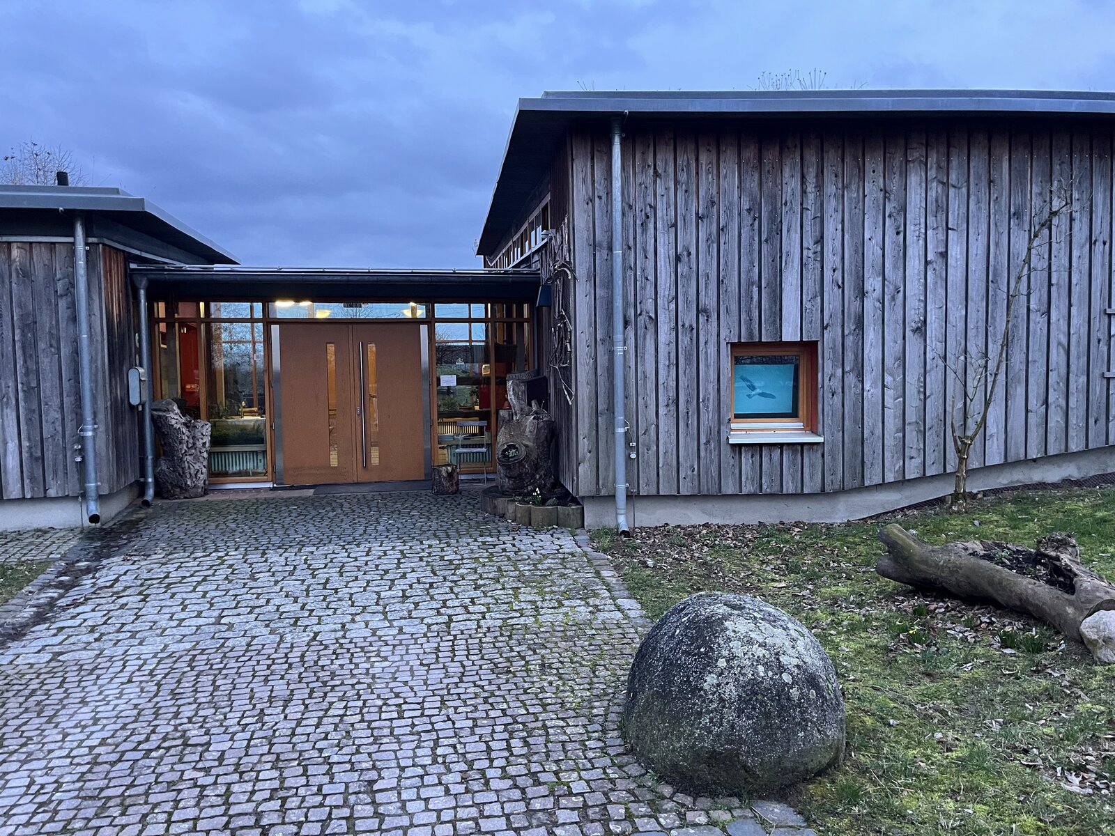 Entrance of the Naturschutzzentrum Bergstraße at dusk, a wooden building with a cobbled path and a large boulder in the foreground
